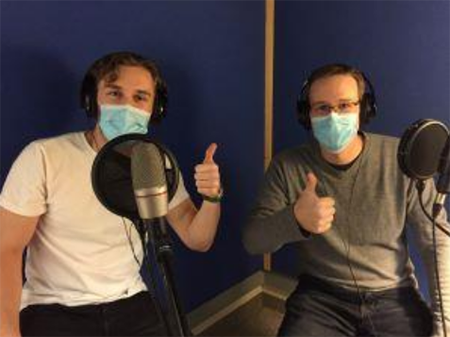Two men wearing masks sit at microphones for a podcast, giving thumbs up