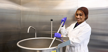 A student in a lab coat and hair net stirs a giant stainless steel cooking pot with a large paddle.