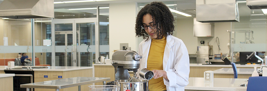 A student adds ingredients to a mixer in a food lab. 