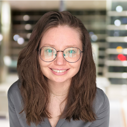 Cait, a white woman with brown shoulder-length hair wears a gray blouse and round, black-rimmed glasses as she smiles into the camera.