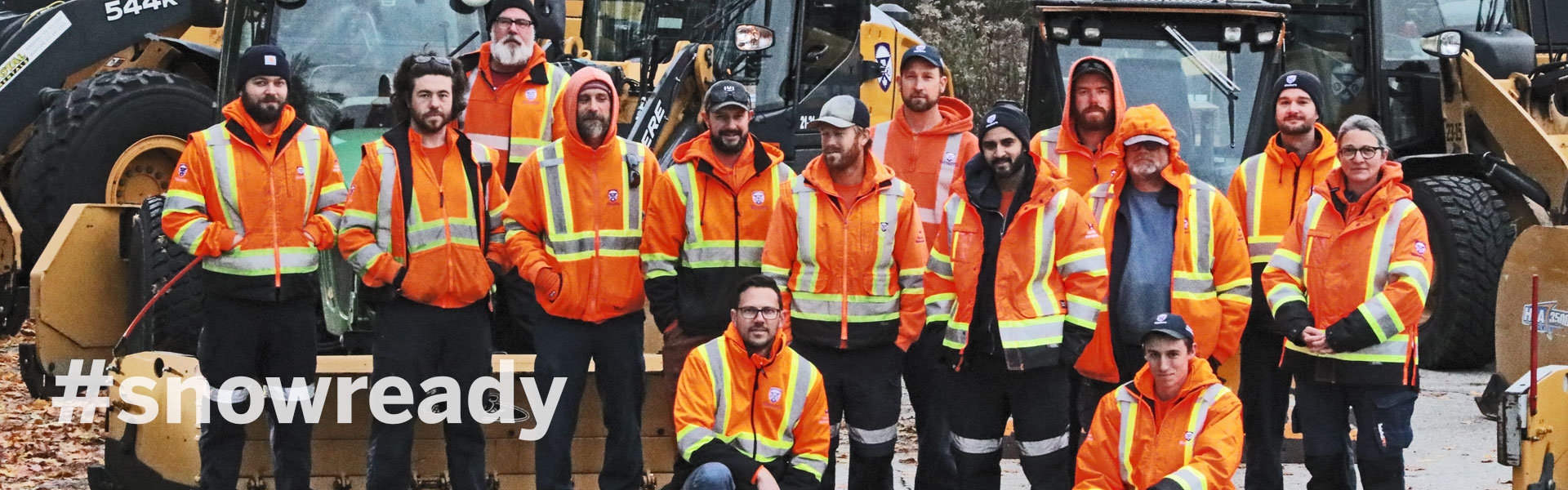 Our snow equipment is tuned up and our operators are prepared for the winter ahead. The image is a group shot of the team, dressed in high visibility vest and posing in front of the snow removal vehicles.