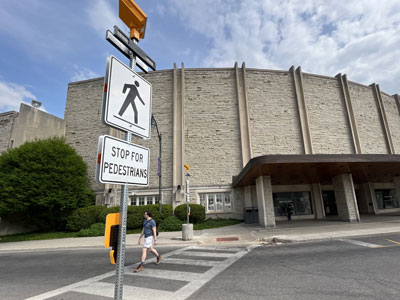 Alumni Circle, with Alumni Hall in the background and a button-activated pedestrian crosswalk in the foreground. A pedestrian is walking across the road.