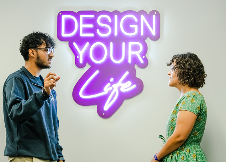 Two people talking with each other between a sign that says design your life
