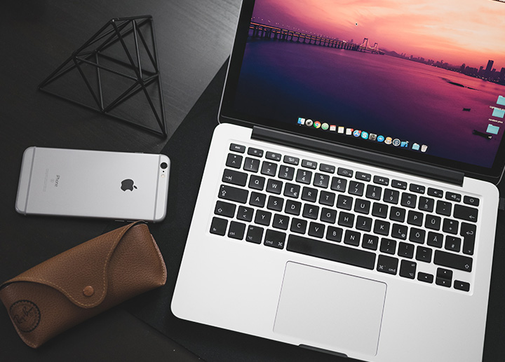 Top view shot of a Mac laptop, Iphone, and Rayban case on a wooden table
