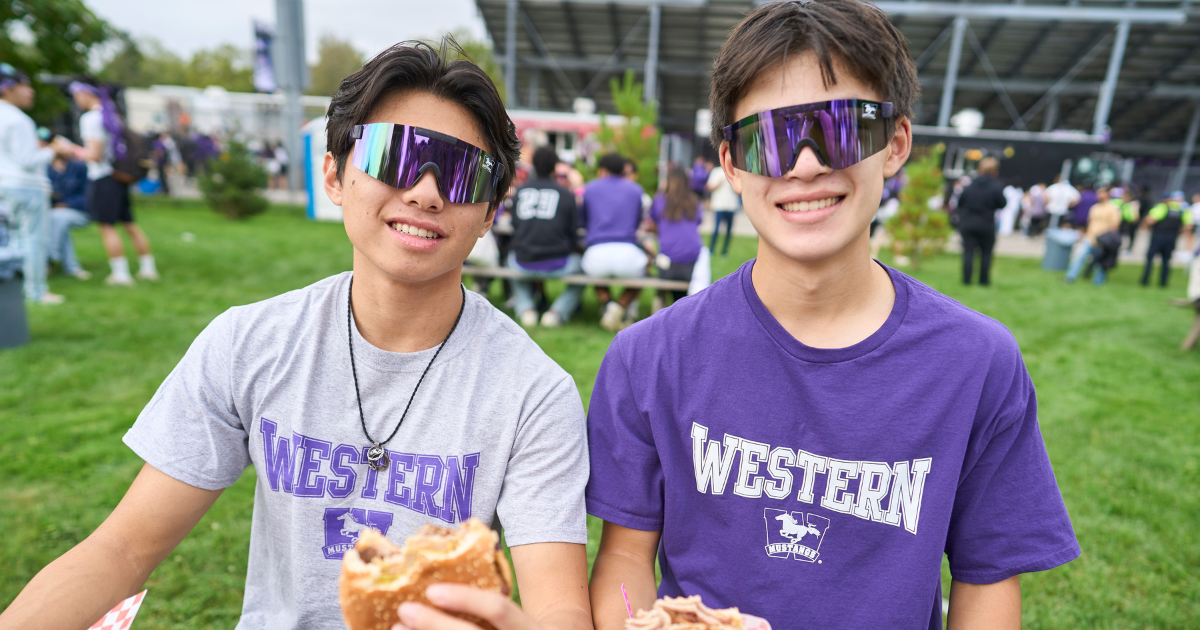 Two students sitting at a picnic table, holding burgers, wearing Western tshirts, smiling for a photo.